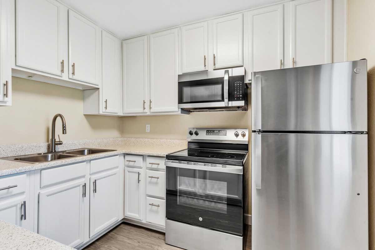 A modern kitchen featuring white cabinets, a stainless steel refrigerator, an oven, and a microwave. The countertop is a light color, and there is a double sink with a sleek faucet. The overall design is bright and contemporary, with a neutral wall color and wood-like flooring.