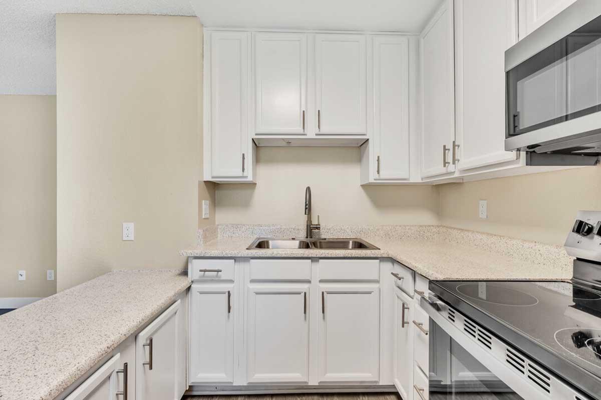 Modern kitchen featuring white cabinetry, a stainless steel sink, and granite countertops. The kitchen includes a cooktop and an overhead microwave, all set against a light beige wall. Brightly lit with an open layout, it offers a clean and contemporary design.