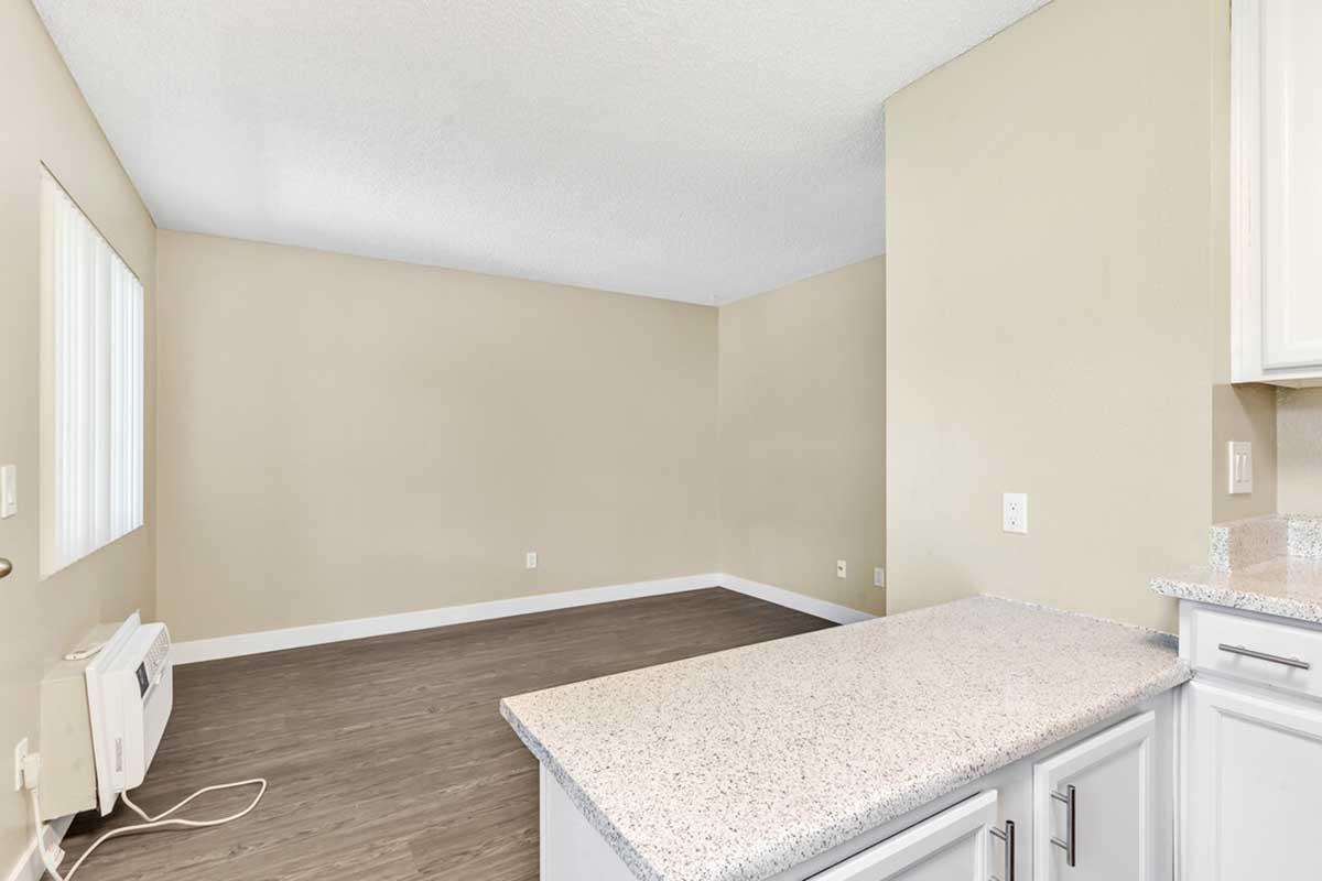A spacious, empty living area with light beige walls and laminate flooring. There is a small kitchen counter on the right, and an air conditioning unit on the left. A window allows natural light to enter the room, which features no furniture or decorations.