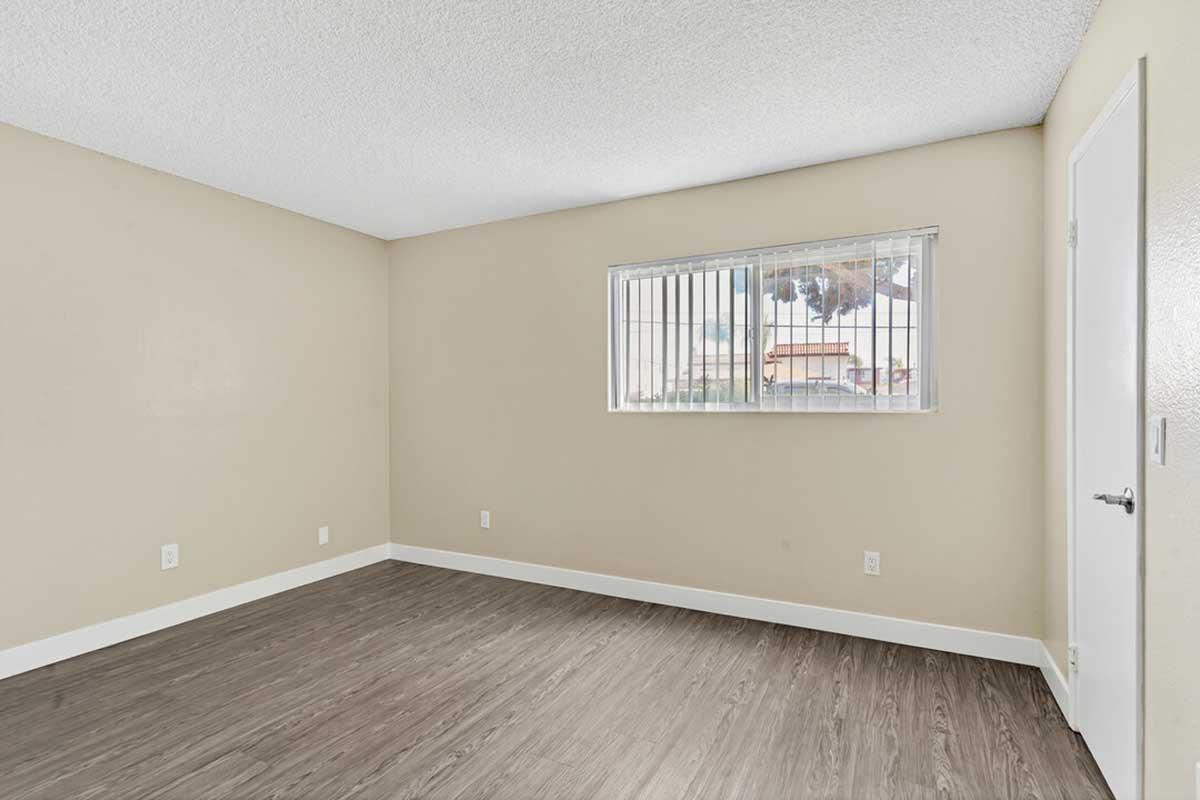 Empty room with beige walls and a textured ceiling. A window with barred security grilles allows natural light to enter. The floor is covered in a light wood-like laminate. There are no furnishings or decorations, creating a spacious and minimalistic atmosphere.