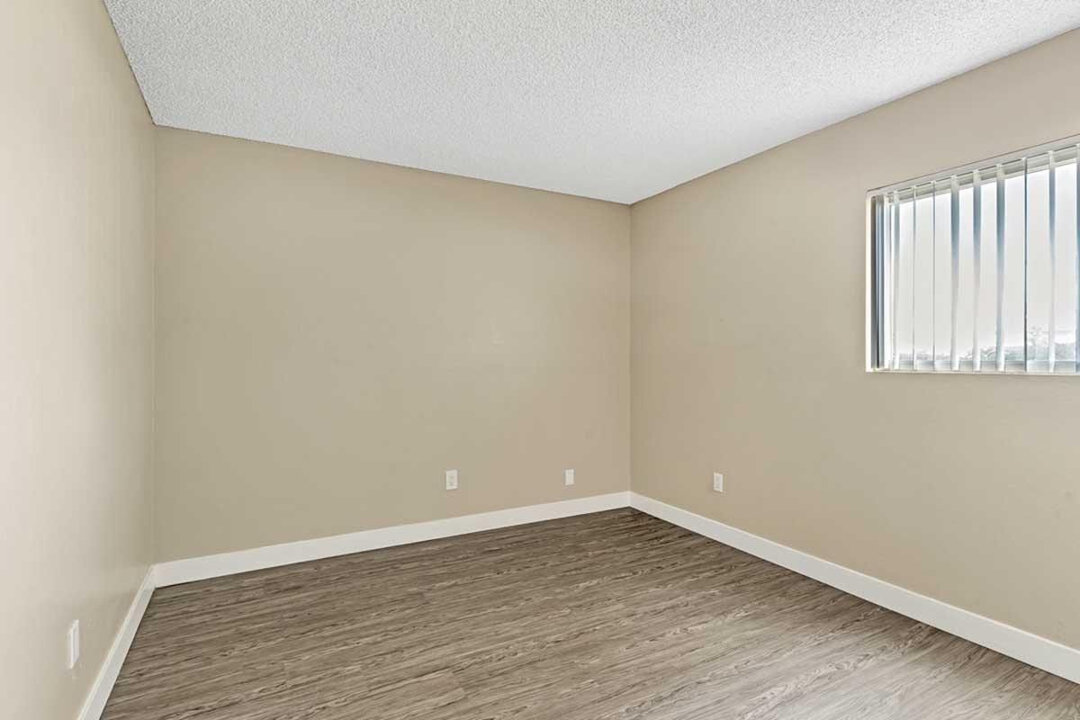 Empty room with light beige walls, a textured ceiling, and a window with vertical blinds allowing natural light. The floor is covered in light wood-like laminate. There are no furnishings or decorations, creating a spacious and minimalistic appearance.