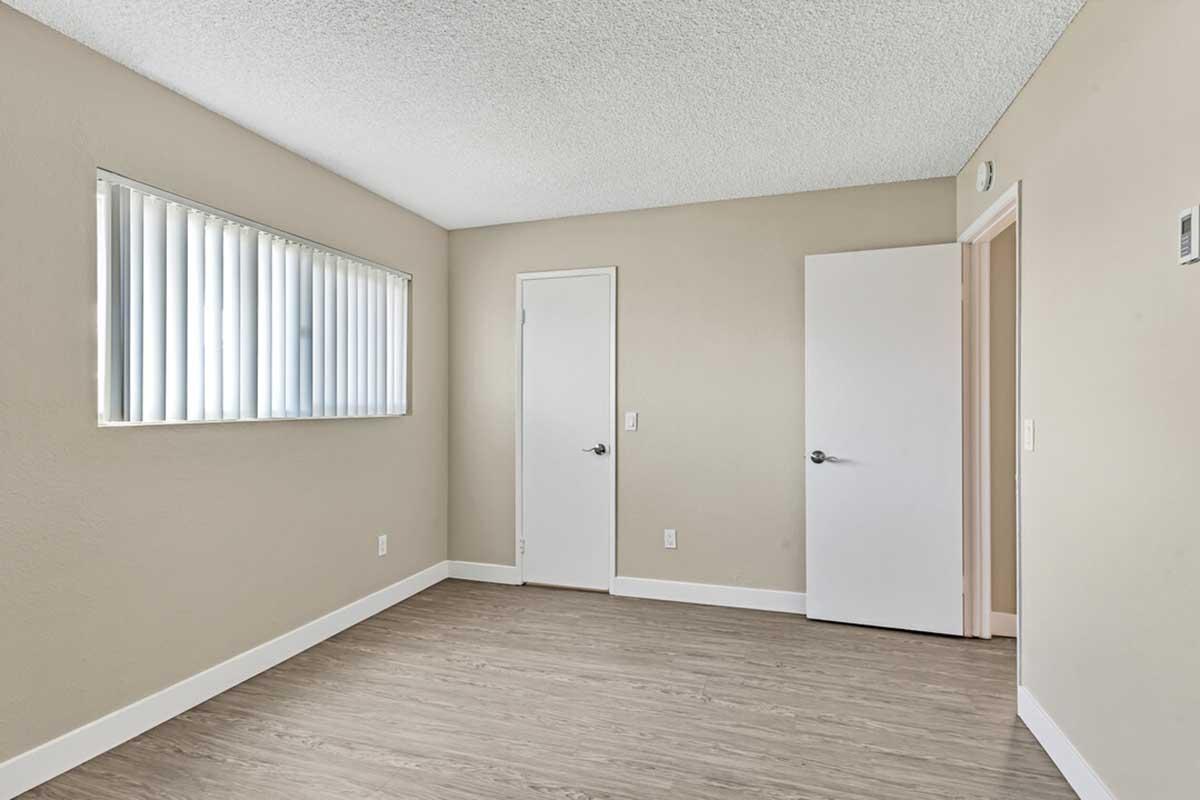 A simple, empty room with light beige walls, a textured ceiling, and laminate flooring. There is a window with vertical blinds on one wall and two doors: one leading to the exterior and the other to an adjoining space. The room is well-lit with natural light.