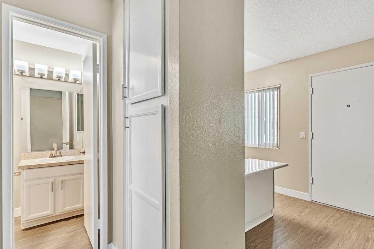 Interior view of a modern bathroom with a vanity and mirror on the left, featuring light-colored cabinetry and countertop. On the right, there is a doorway leading to a bright room with a window covered by vertical blinds. The walls are painted in neutral tones, and the floor is a warm wood finish.