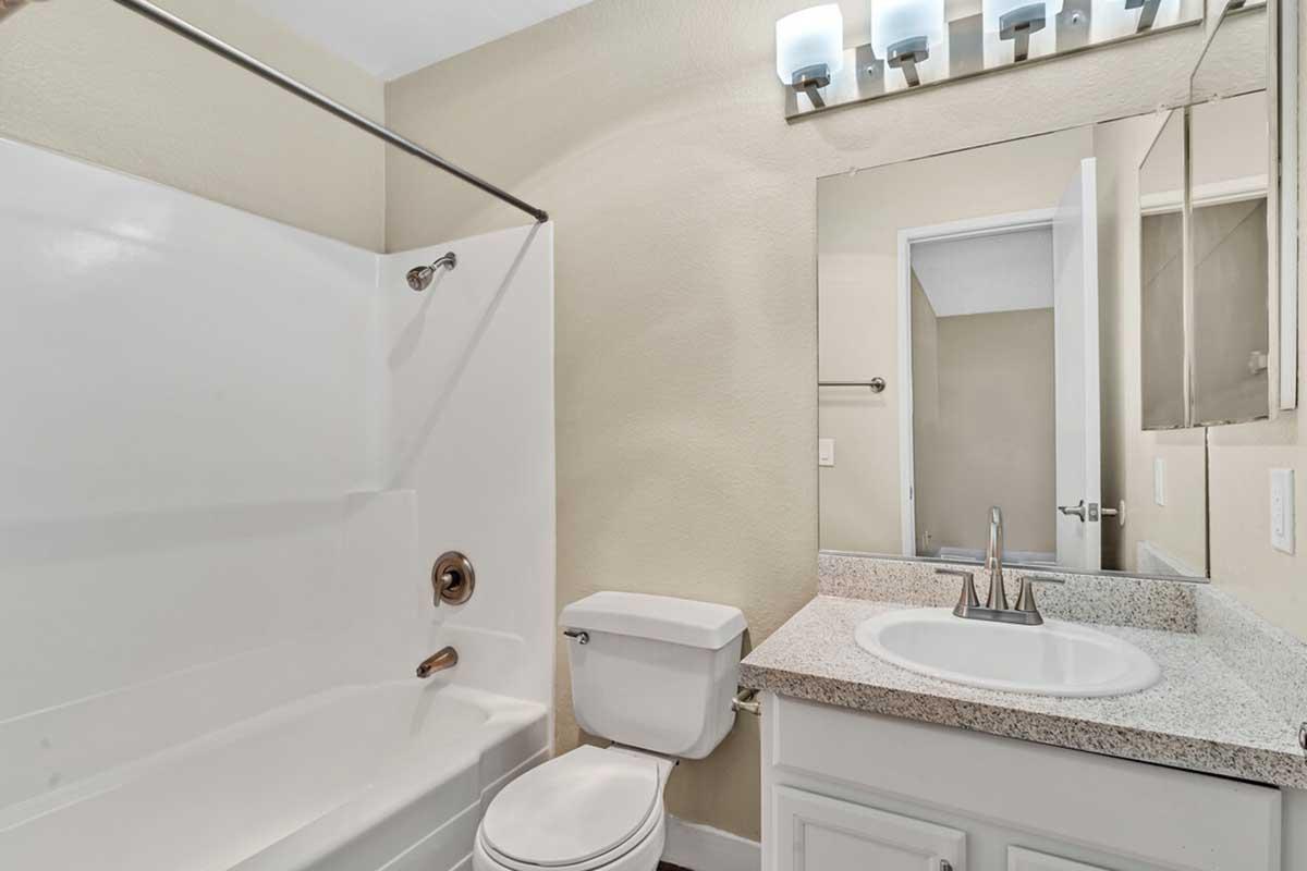 A clean and modern bathroom featuring a bathtub with a shower, a white toilet, and a granite countertop sink. The walls are painted in a light beige color, and there is a mirror above the sink with two light fixtures. A door leads to another room, enhancing the space's functionality.