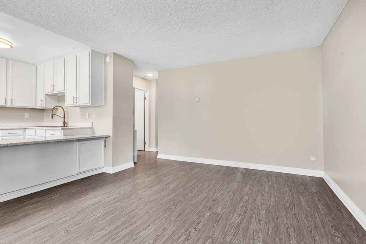 A spacious, empty living room with light-colored walls and wood-like flooring. The kitchen is partially visible on the left, featuring light cabinetry and a modern faucet. A doorway leads to another room in the background, providing a sense of openness and potential for decoration.