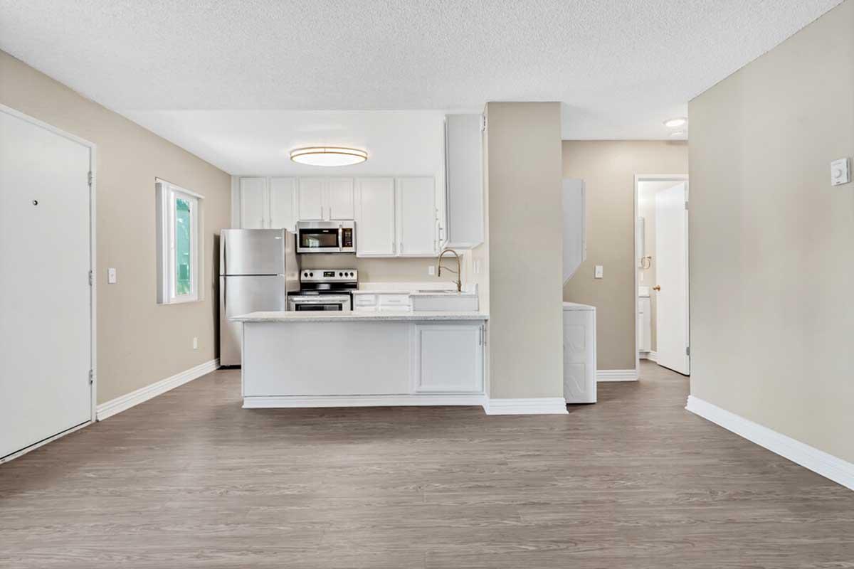 A modern, airy kitchen and living space with light-colored walls and wood-style flooring. Features include a stainless steel refrigerator, oven, and microwave, along with white cabinetry. A doorway leads to a small hallway, and a window allows natural light into the area.