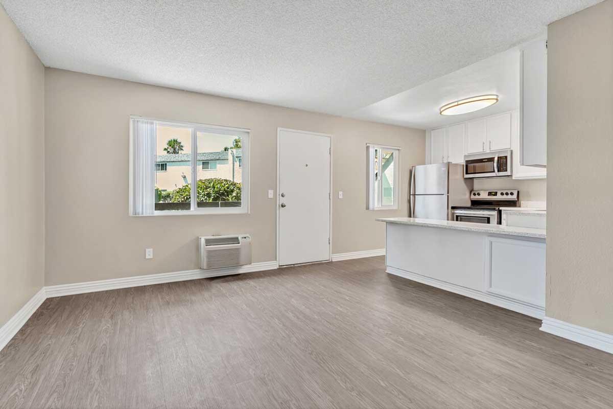 A modern, unfurnished living room with light brown walls and large windows. The space features a white kitchen with sleek appliances, a door leading outside, and an air conditioning unit. Natural light illuminates the room, highlighting the laminate flooring.
