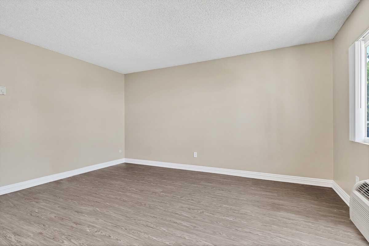 A spacious, empty room with light beige walls and a wooden floor. There is a window on the right side allowing natural light to enter, and the overall atmosphere is bright and open, suitable for various uses or decoration. No furniture is present.
