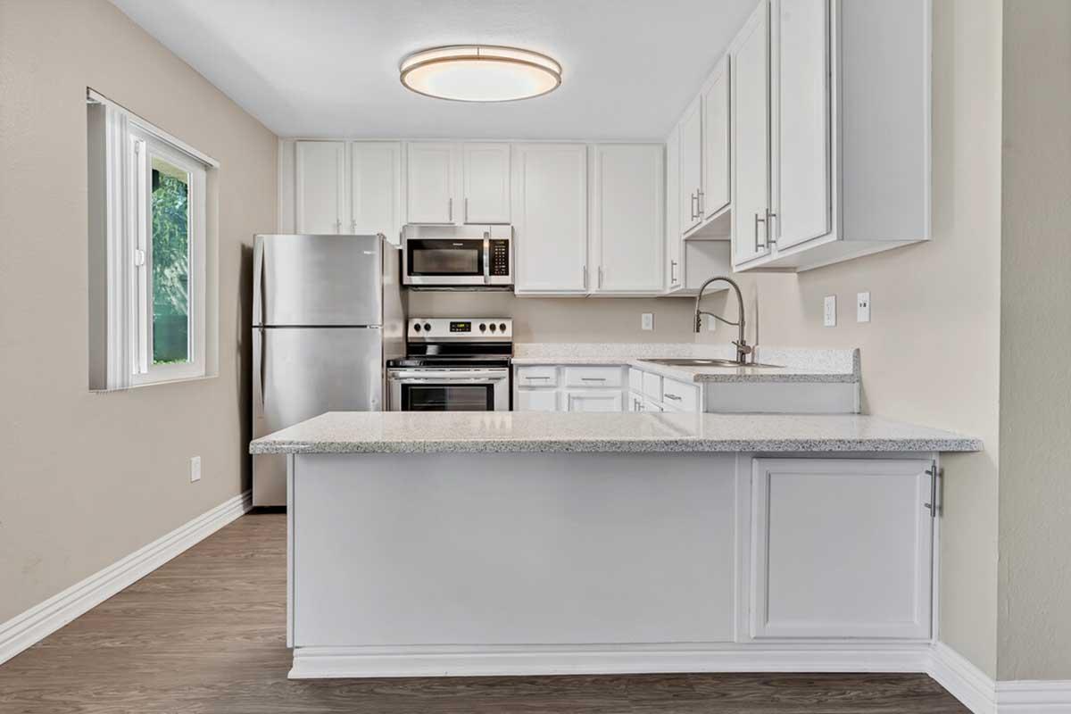 Modern kitchen with white cabinets, stainless steel appliances including a refrigerator and microwave, and a granite countertop. The space features an open layout with a view of a window and natural light, along with a circular ceiling light fixture. The flooring is a light wood finish.