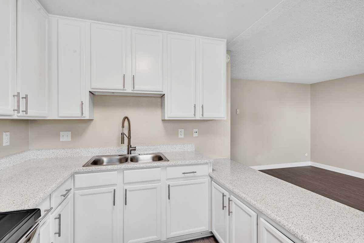 Modern kitchen with white cabinets and a spacious granite countertop featuring a double sink. The design is open and bright, with a neutral-colored wall in the background and a glimpse of a living area with wooden flooring.