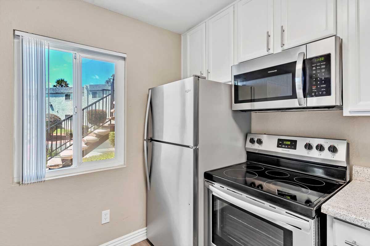 A modern kitchen featuring stainless steel appliances including a refrigerator, microwave, and oven. There is a window with sheer blinds providing natural light and a view of a staircase outside. The cabinetry is white and the countertop has a light granite finish.