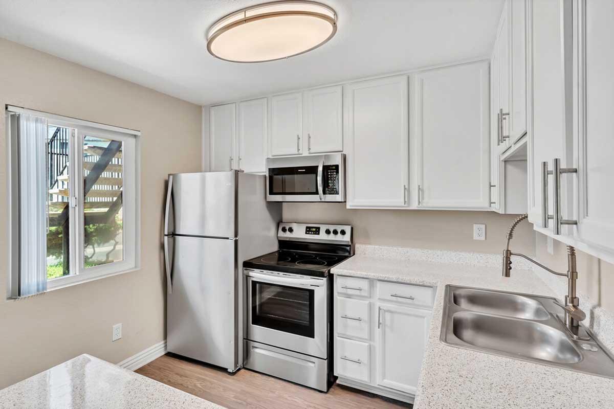 Modern kitchen featuring white cabinetry, stainless steel appliances including a refrigerator, microwave, and oven. The countertop is light-colored and there are dual sinks. A window lets in natural light, showing part of an outdoor space. The kitchen has a clean and contemporary design.
