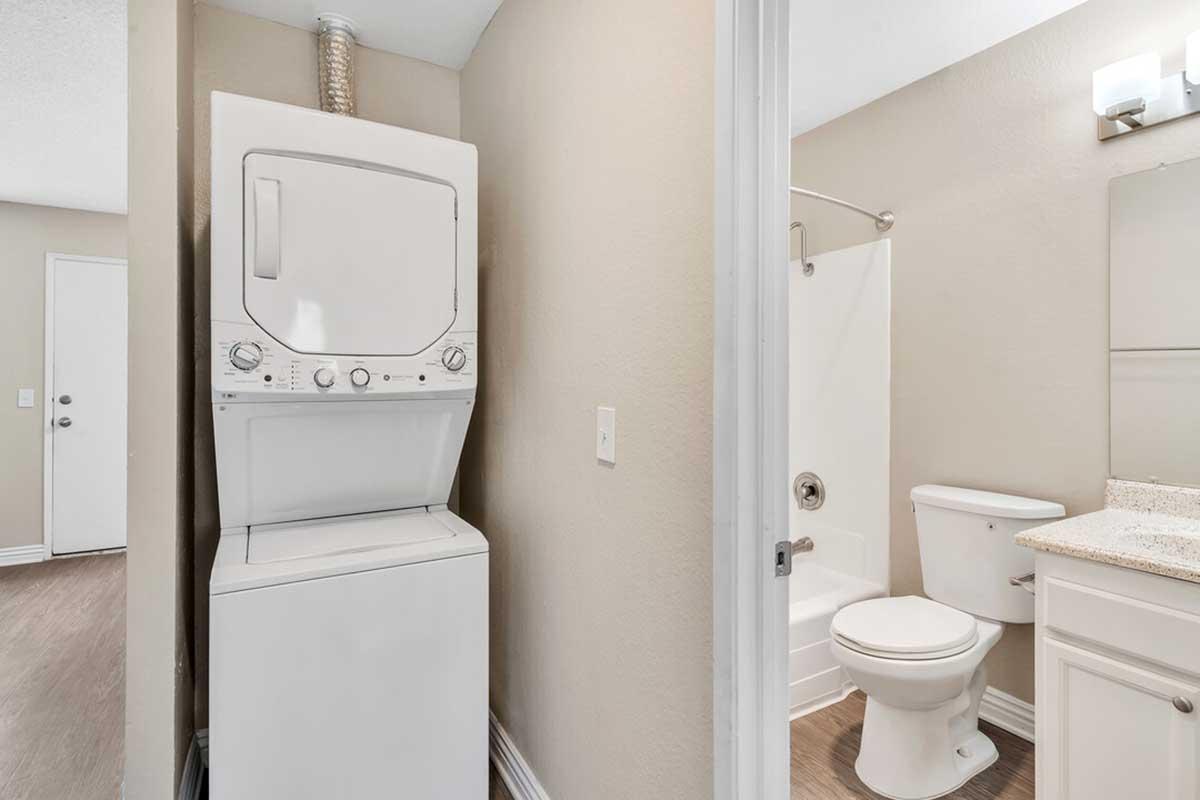 A small laundry area featuring a stacked washer and dryer next to a bathroom. The bathroom includes a shower stall, toilet, and a vanity with a mirror. Light-colored walls and flooring create a bright, airy atmosphere. A door leads to another room.