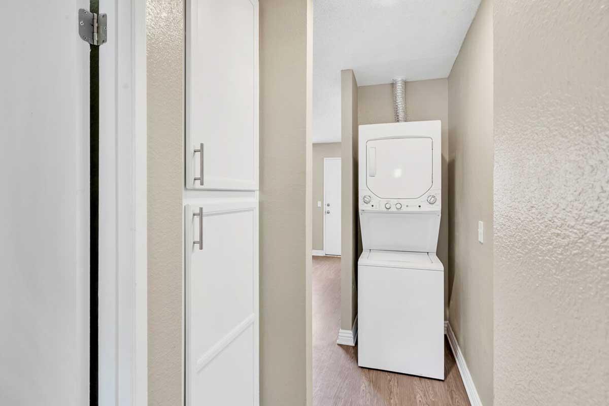 A narrow hallway featuring a stackable washer and dryer on the right. Light beige walls create a warm atmosphere, and there are doors leading to other rooms in the background. The floor is wooden, adding a homey touch to the space.