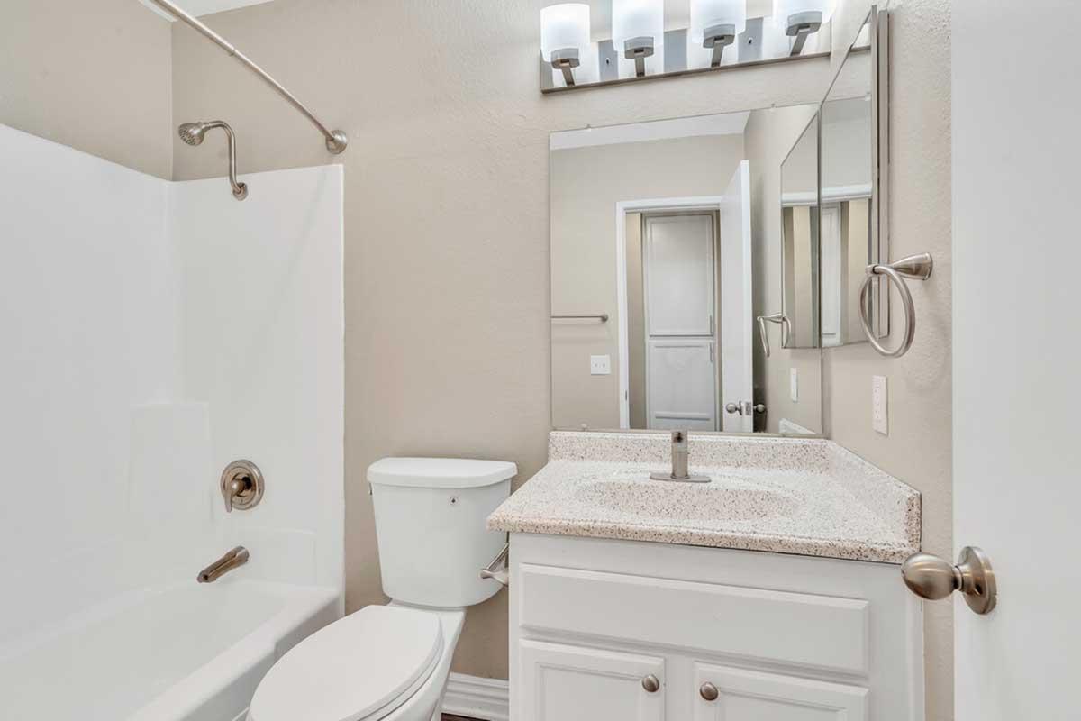 A clean and modern bathroom featuring a white bathtub with shower, a sink with a granite countertop, a white toilet, and a large mirror. The walls are painted in a neutral color, and there are light fixtures above the mirror. A door leads to another room in the background.