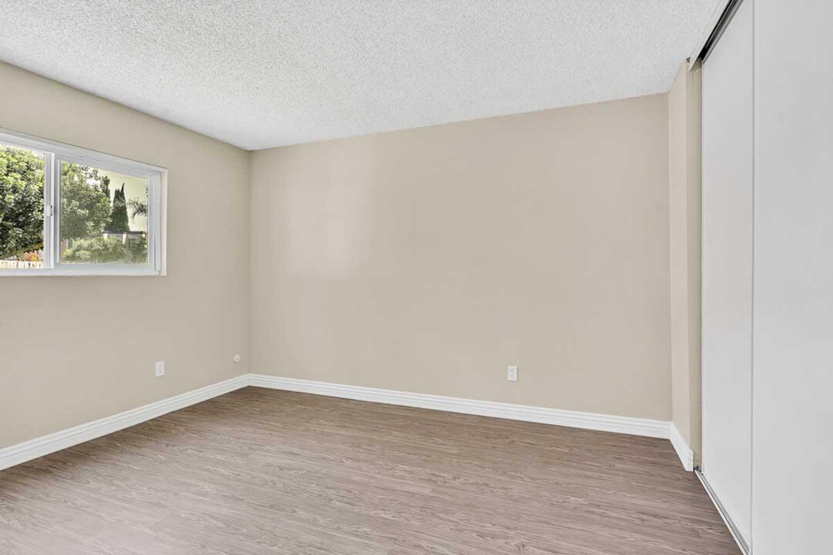 A vacant, light-colored room with textured walls and a window allowing natural light. It features a smooth, brown laminate floor and white baseboards. There is a closed sliding door on one side, and the space appears clean and minimalistic, suitable for various interior design styles.