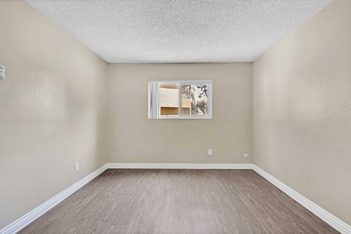 Empty room with light beige walls and a textured ceiling. A small window on one wall allows natural light to enter. The floor is covered with light-colored laminate, enhancing the spacious feel of the area. No furniture or decorations are present.