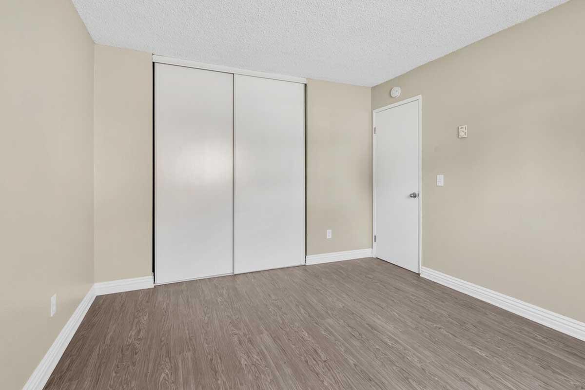 Empty room with light brown walls, a textured ceiling, and laminate flooring. The room features a closed sliding door closet and a white door on one side. There is minimal decoration, creating a spacious and neutral atmosphere. Natural light may be present, enhancing the bright appearance of the space.