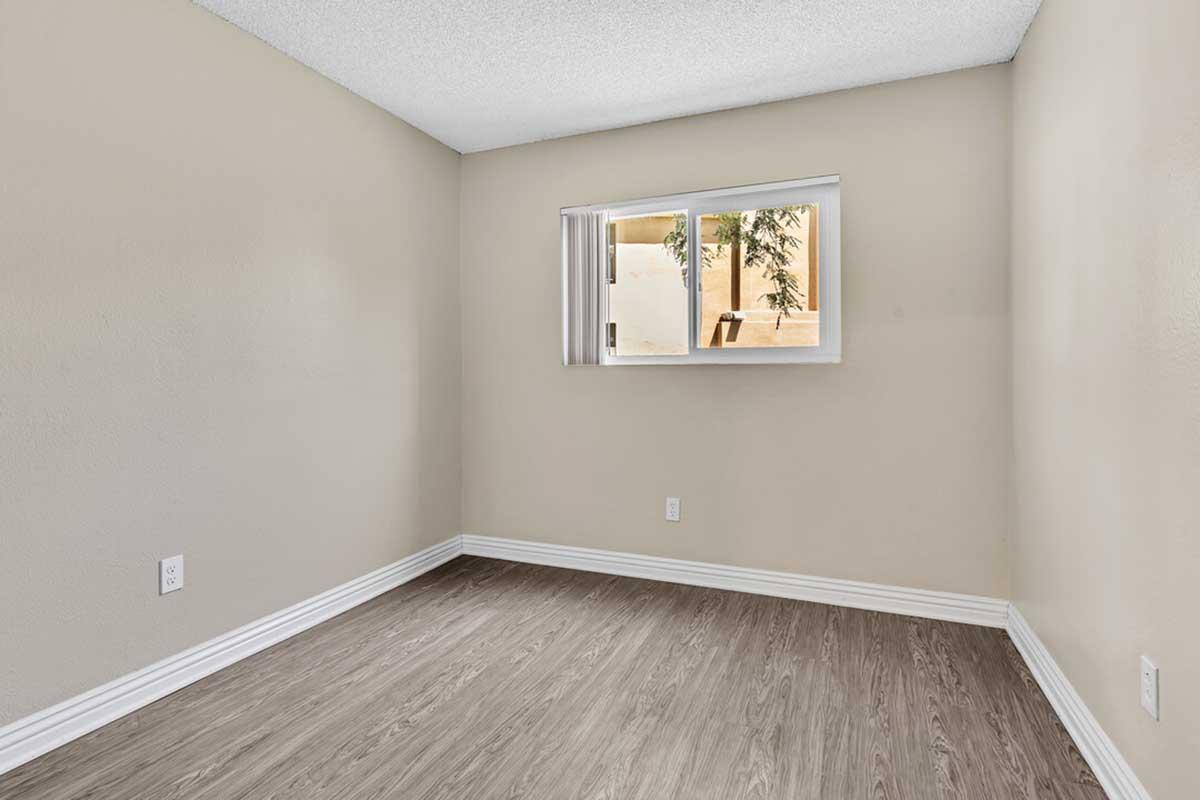 Empty room with beige walls and a window on one side. The flooring appears to be a light-colored laminate. There are no furnishings or decorations, creating a minimalist and spacious feel. Natural light enters through the window, highlighting the simplicity of the space.