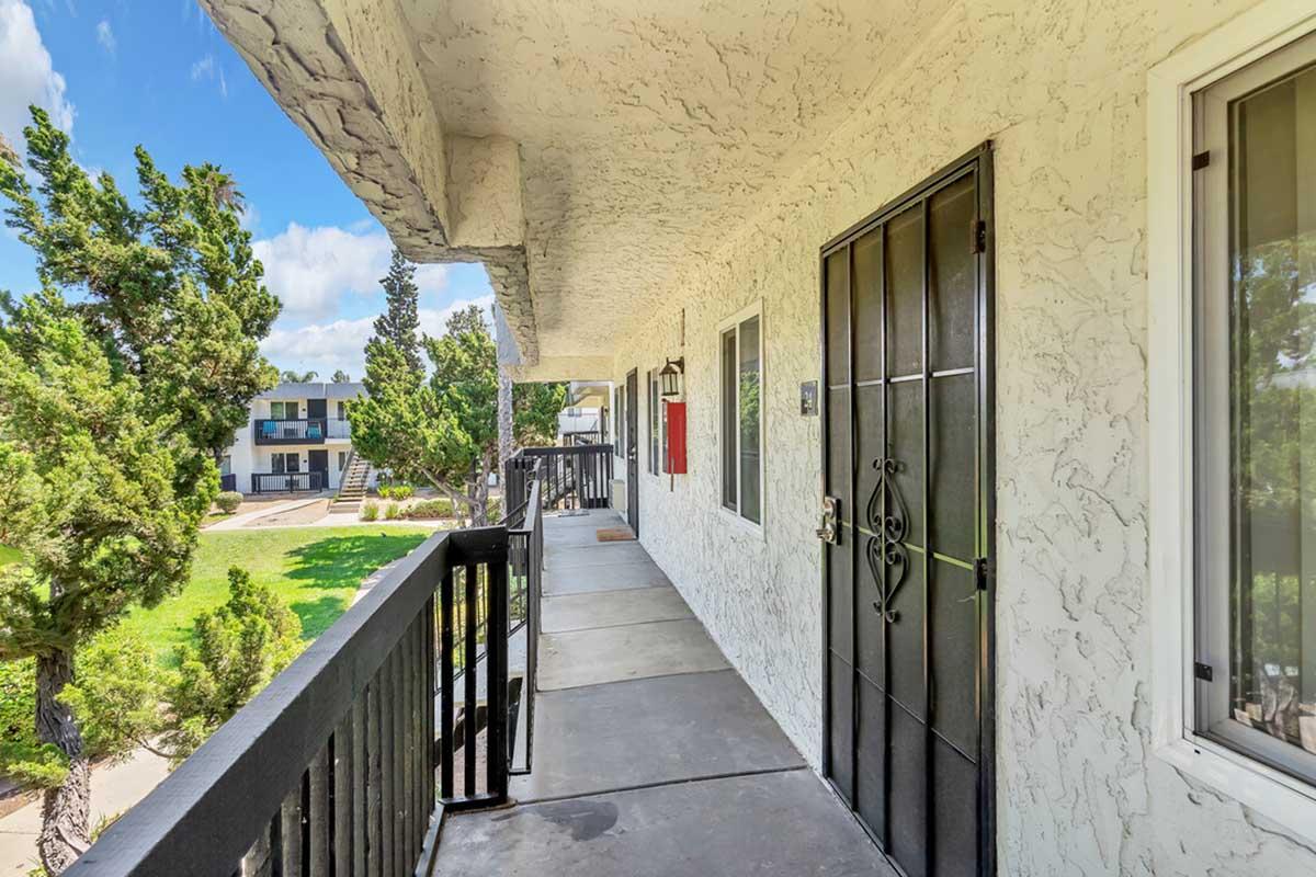 A view of a concrete hallway with a black door on the left, surrounded by a white textured wall. Green trees are visible in the background, along with a grassy area and a glimpse of other buildings. The scene is bright and sunny, suggesting a pleasant outdoor environment.