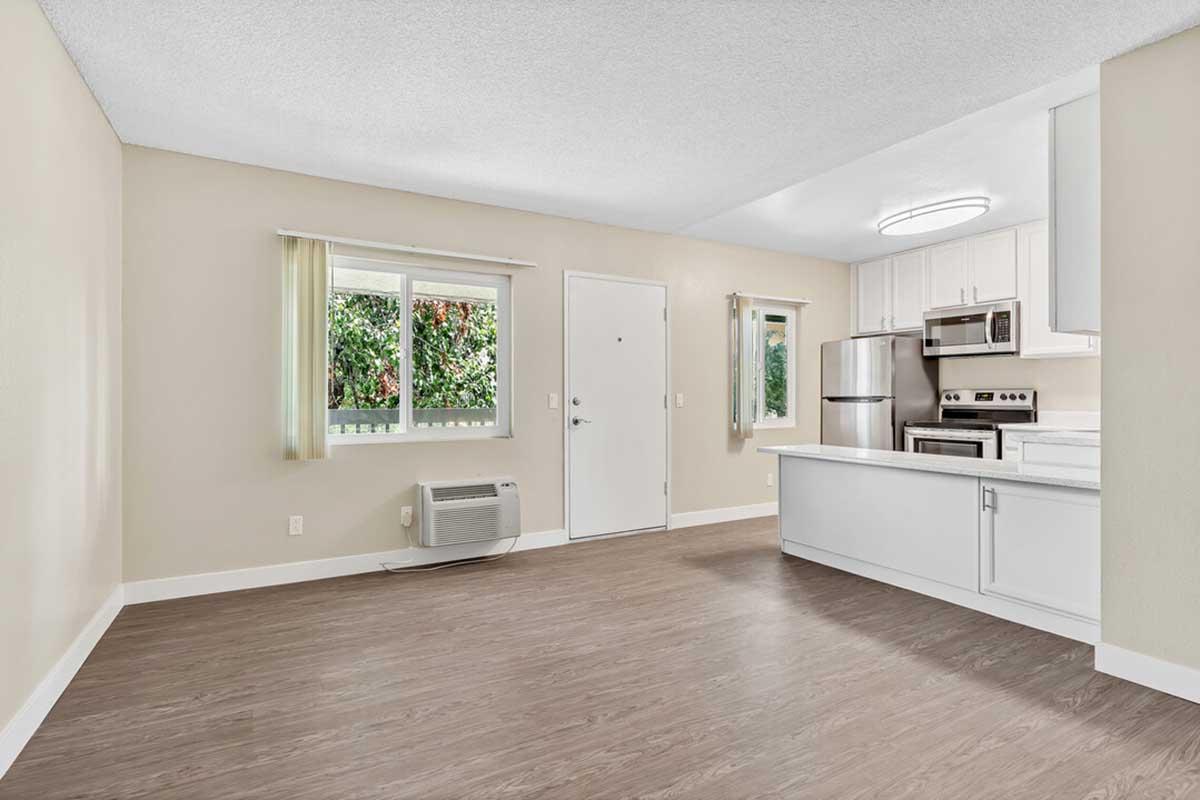 A modern, vacant kitchen and living area featuring light-colored walls, a large window with green outside views, and an air conditioning unit on the floor. The kitchen includes stainless steel appliances, white cabinetry, and a small island. The flooring is a light wood laminate.