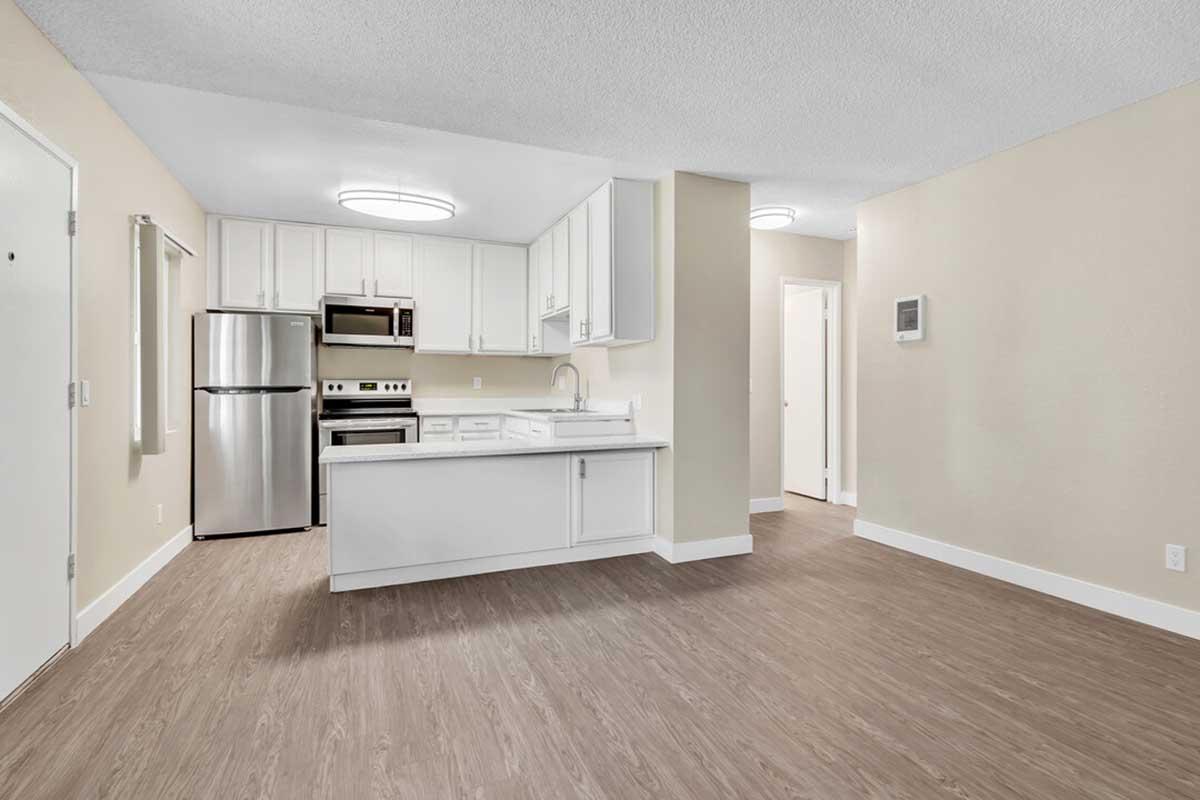 Modern kitchen and living area featuring white cabinetry, stainless steel appliances, and wood-style flooring. The kitchen island provides additional counter space, while large overhead lights enhance the bright atmosphere. A closed door leads to a room or hallway, adding to the open layout.
