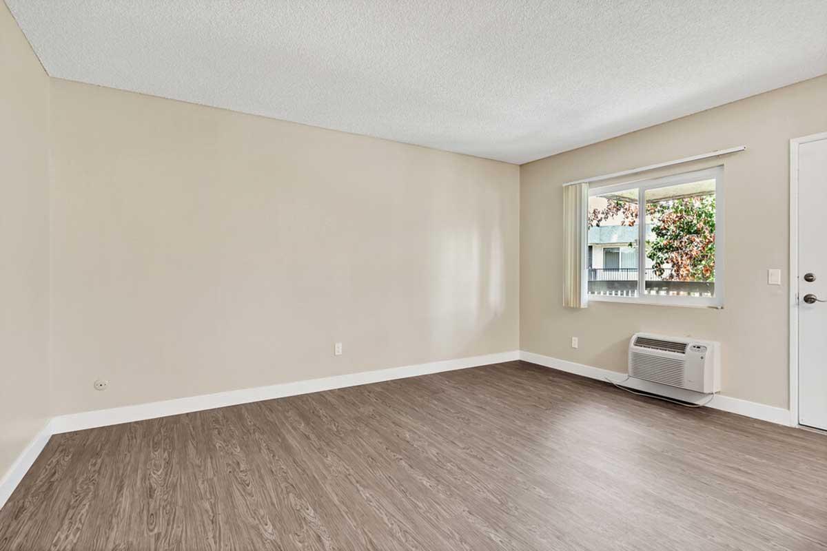 A vacant room with a light beige wall, a window with sheer curtains, and a wall-mounted air conditioning unit. The floor features tan laminate flooring, and there is a door on the right side, leading outside. The space is well-lit and appears modern and minimalist.