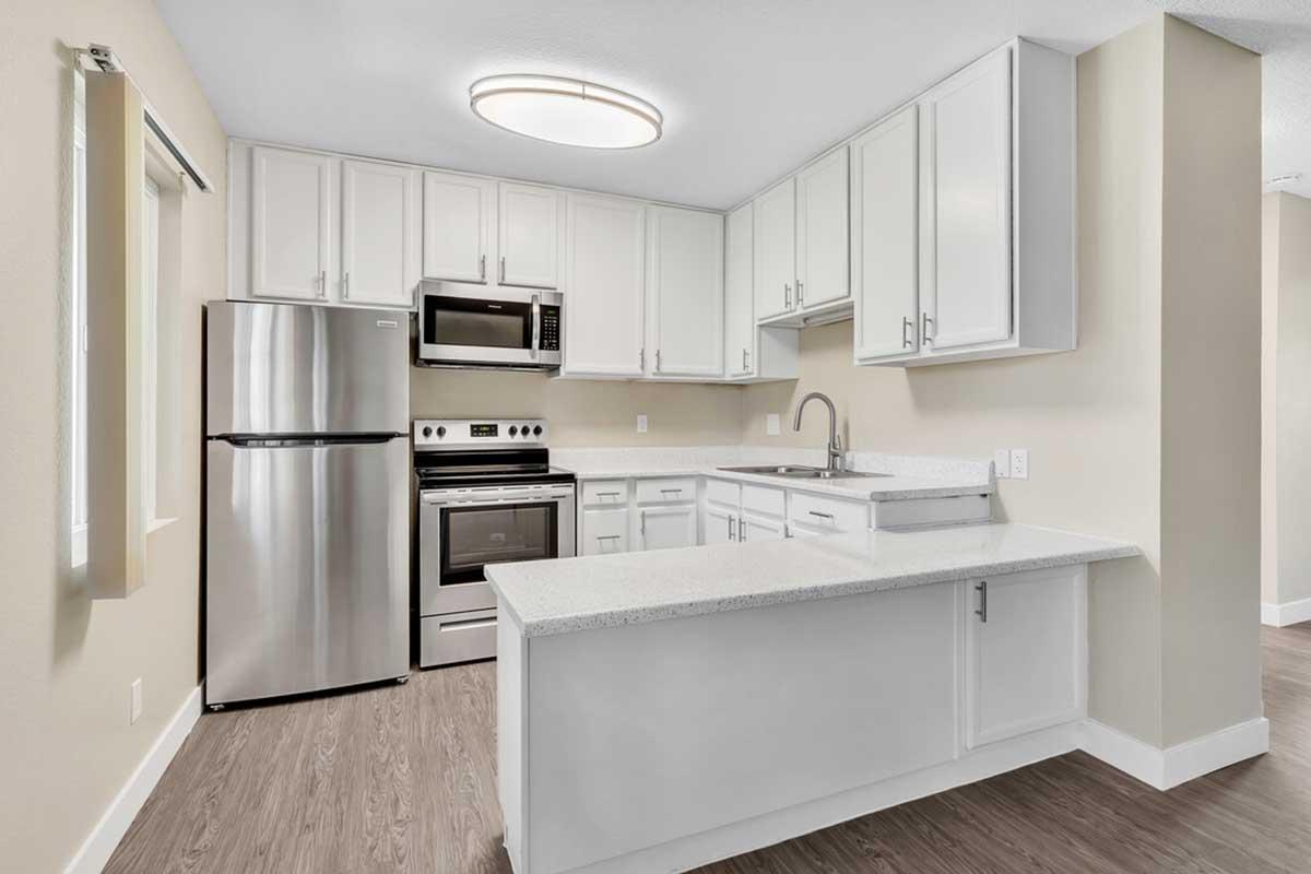 Modern kitchen featuring white cabinetry, stainless steel appliances (refrigerator, microwave, oven), and a light-colored countertop. The space has a contemporary design with natural wood flooring and ample lighting from a ceiling fixture.