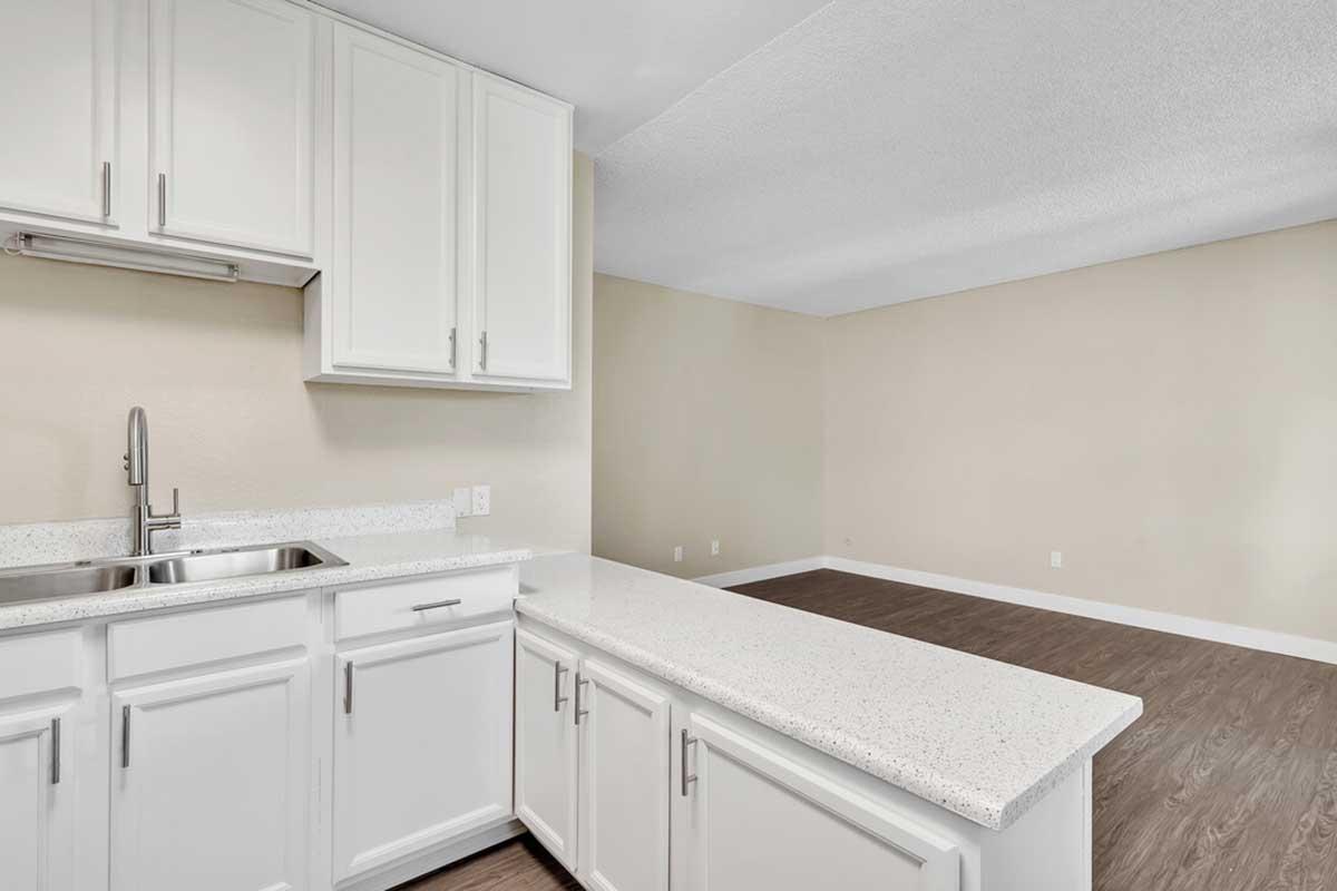 Modern kitchen with white cabinets and a granite countertop, featuring a stainless steel sink and faucet. The adjacent living area has neutral-colored walls and brown laminate flooring, creating a spacious and open feel.