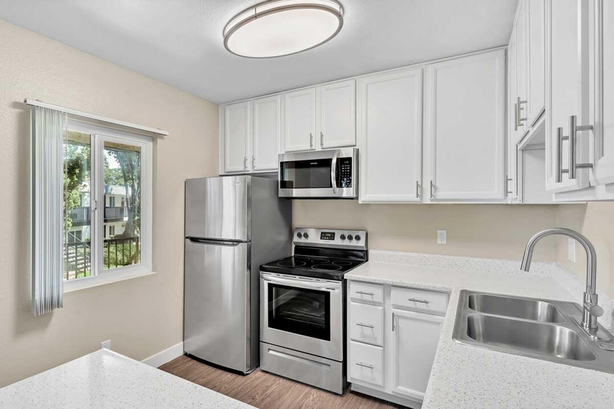 Modern kitchen with white cabinetry, stainless steel appliances including a refrigerator, microwave, and oven. Features a double sink and a window providing natural light, with light-colored walls and a simple ceiling light fixture. The countertops are white and speckled.