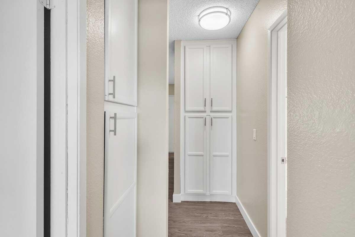 A well-lit hallway with light-colored walls and a brown wooden floor. On the left, there is a closed white door, and on the right, a tall white cabinet with multiple doors. A ceiling light fixture is visible above, enhancing the bright and airy feel of the space.