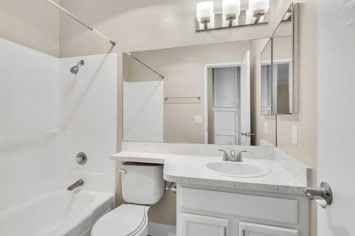 A clean and modern bathroom featuring a white bathtub with a shower, a vanity with a sink, a toilet, and a mirrored cabinet above the sink. Soft neutral walls and bright lighting create an airy ambiance. The space is organized with a towel rack and a door leading to another room.