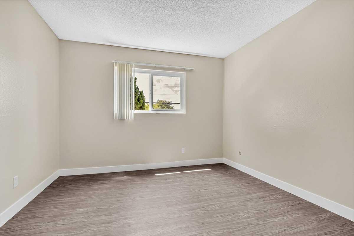 A vacant room with light beige walls and a large window. The floor is a wood-like laminate, and there are white baseboards. A sheer curtain is partially drawn, allowing natural light to enter. The space is empty, creating a minimalist and inviting atmosphere.