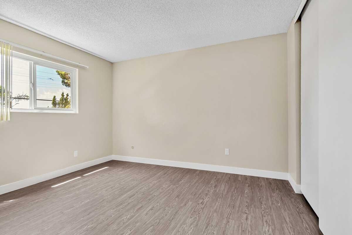 Empty room with light-colored walls and wood-like flooring. A large window on one wall lets in natural light, and there are vertical blinds partially closed. The space appears spacious and well-lit, suitable for various uses.