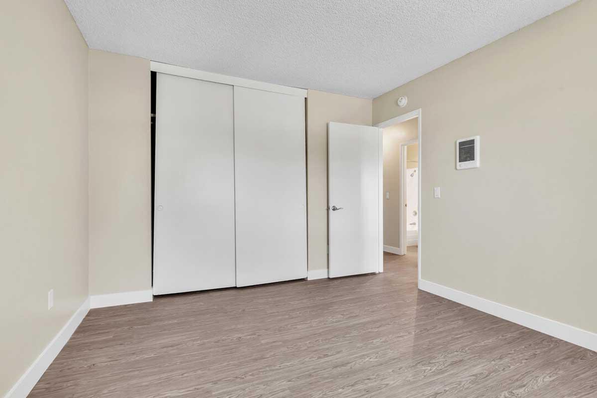 A well-lit, empty room with light beige walls and a wood-like floor. There is a large white closet with sliding doors on one side and a partially open door leading to a bathroom. The room is spacious, featuring modern design elements and minimal decor.