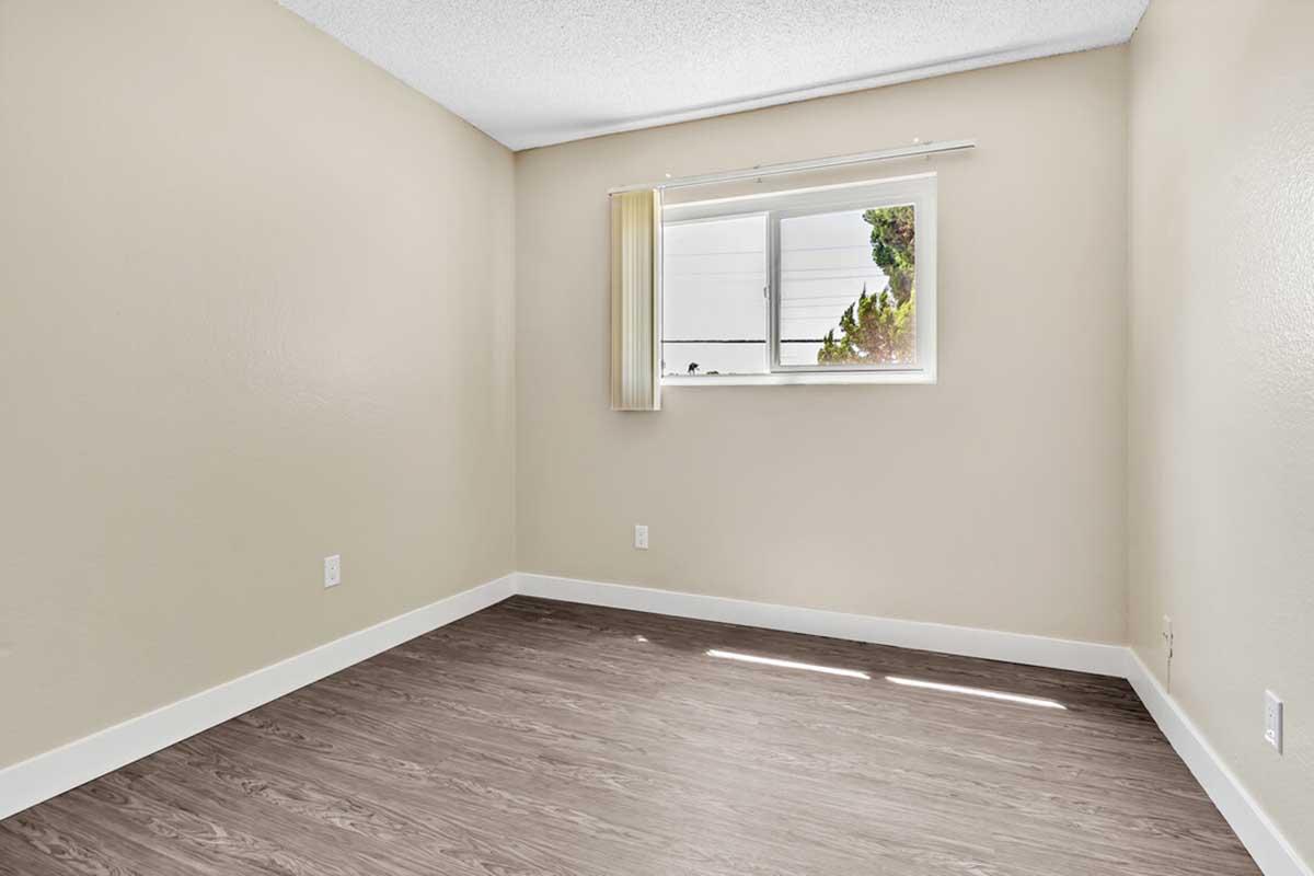Empty room with light beige walls and a window on one side, letting in natural light. The floor features a wood-like laminate, and there are no furnishings or decorations. A sheer curtain partially covers the window, and the overall atmosphere is calm and minimalistic.