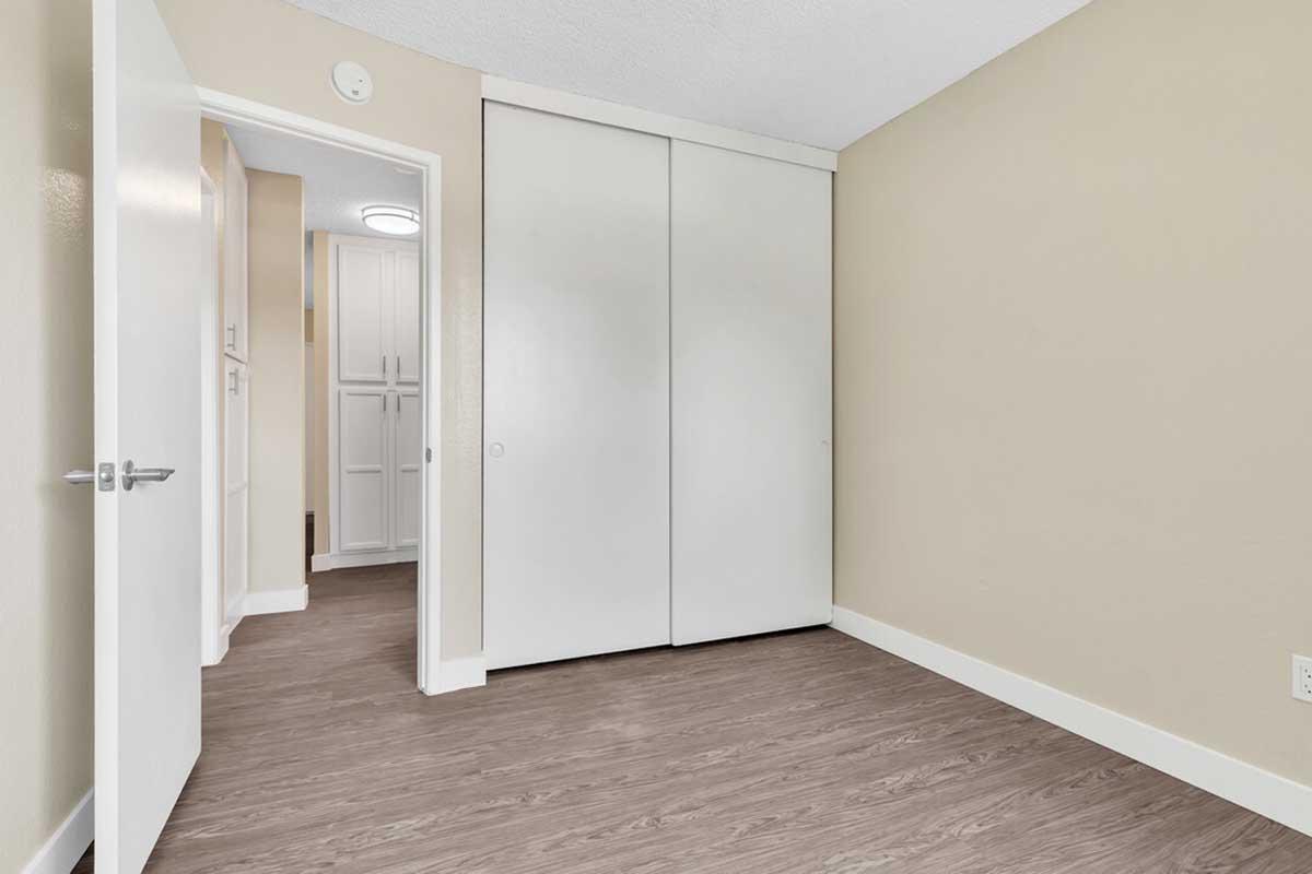 Interior view of a room featuring a light-colored wall and wooden flooring. There is a closed door to the left leading to another space, and a white closet with sliding doors on the right. The lighting is bright, with a ceiling light fixture visible. The overall aesthetic is clean and modern.