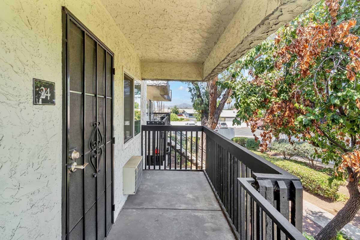 View of a balcony with black railings and a textured wall. The entrance features a secure door with decorative metalwork. Surrounding greenery is visible, along with a glimpse of the street below. Sunlight illuminates the area, creating a welcoming atmosphere.