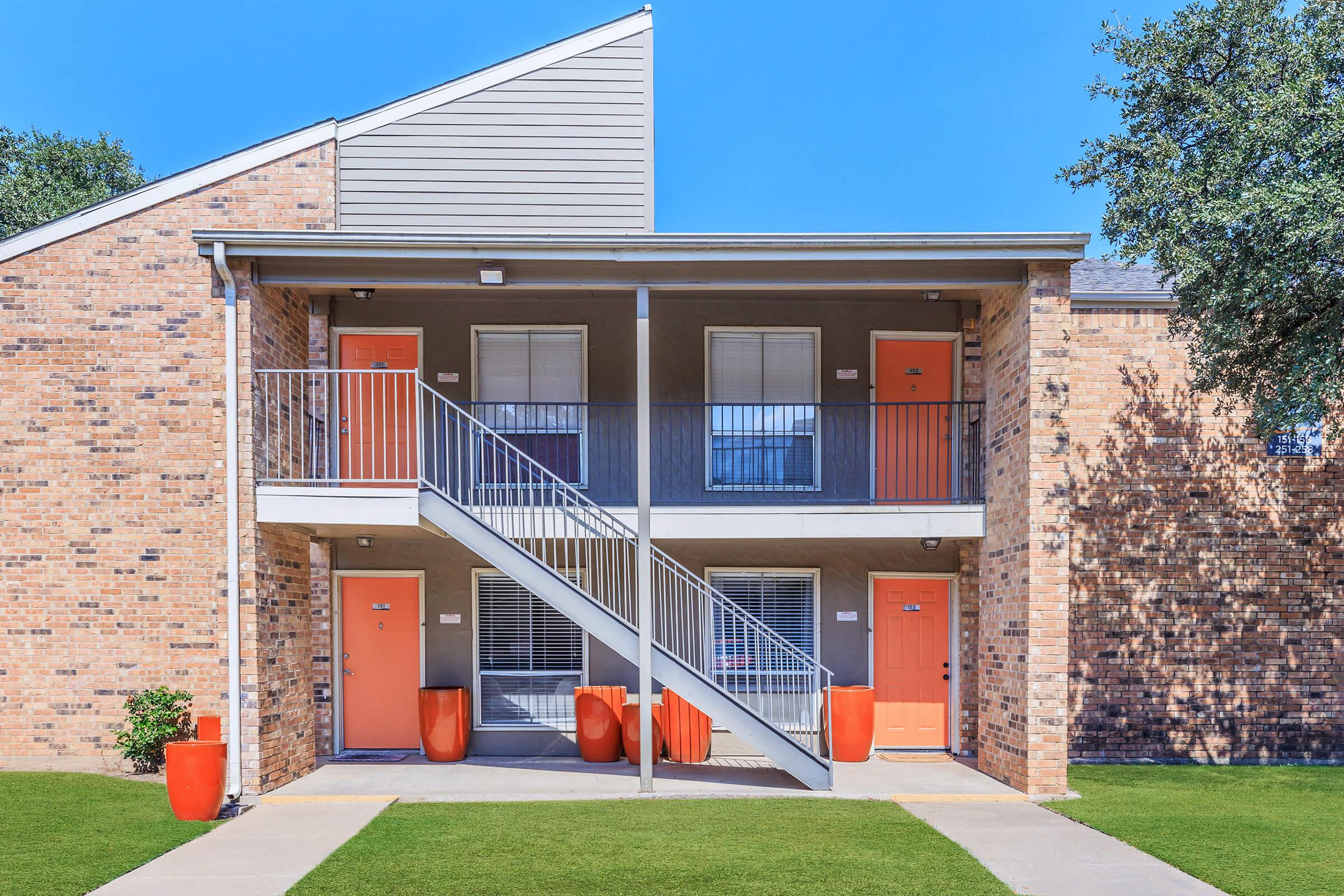 Two-story apartment building with a brick exterior. The entrance features a central staircase leading to the upper level. There are four doors painted in bright orange, with two large orange planters near the stairs. The lawn is well-maintained, and the sky is clear blue, creating a vibrant and inviting atmosphere.