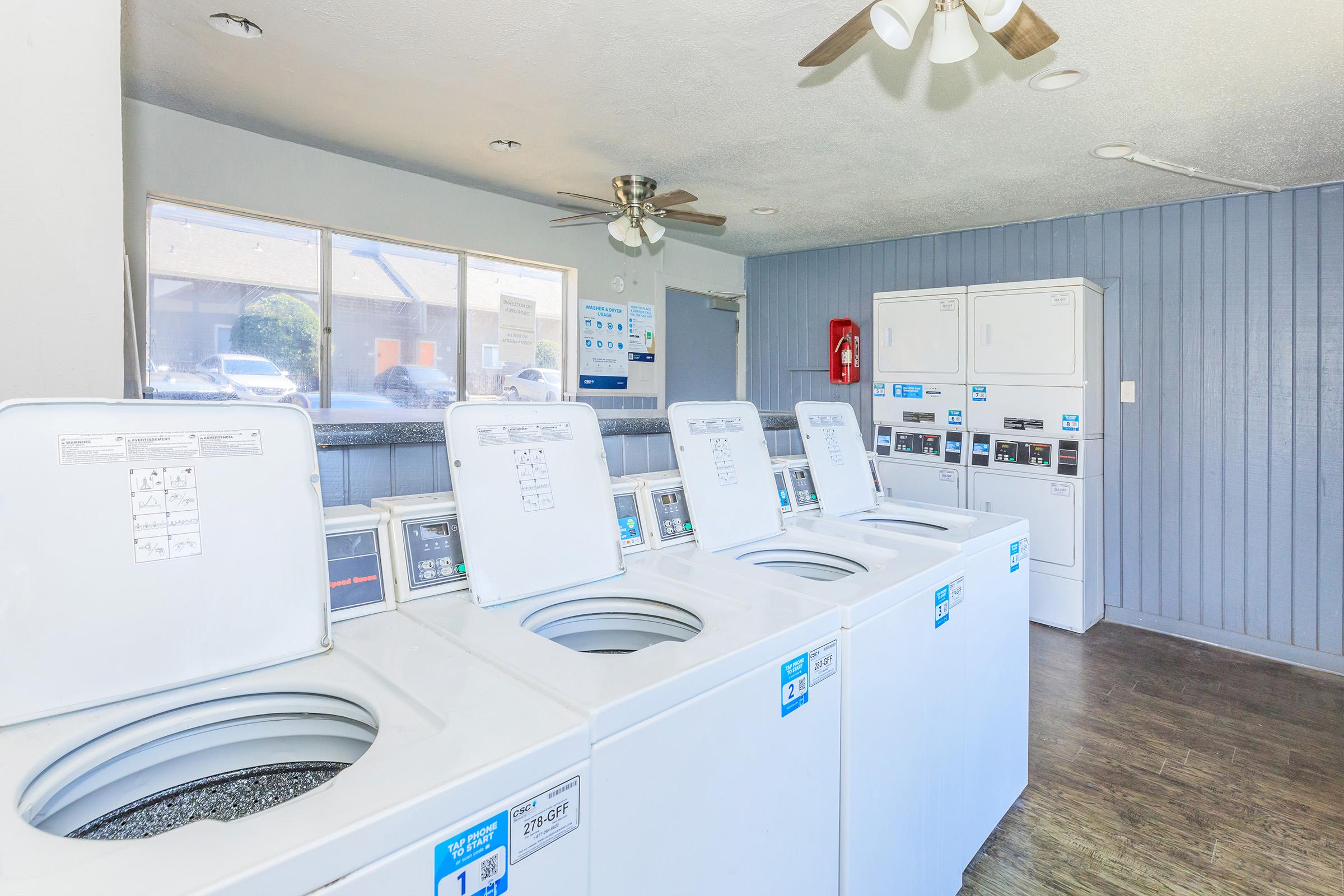 A clean laundry room featuring several rows of washing machines with clearly labeled controls. There’s a ceiling fan, bright lighting, and a window allowing natural light in. Nearby, a fire extinguisher is visible on the wall, contributing to the safety features of the space.