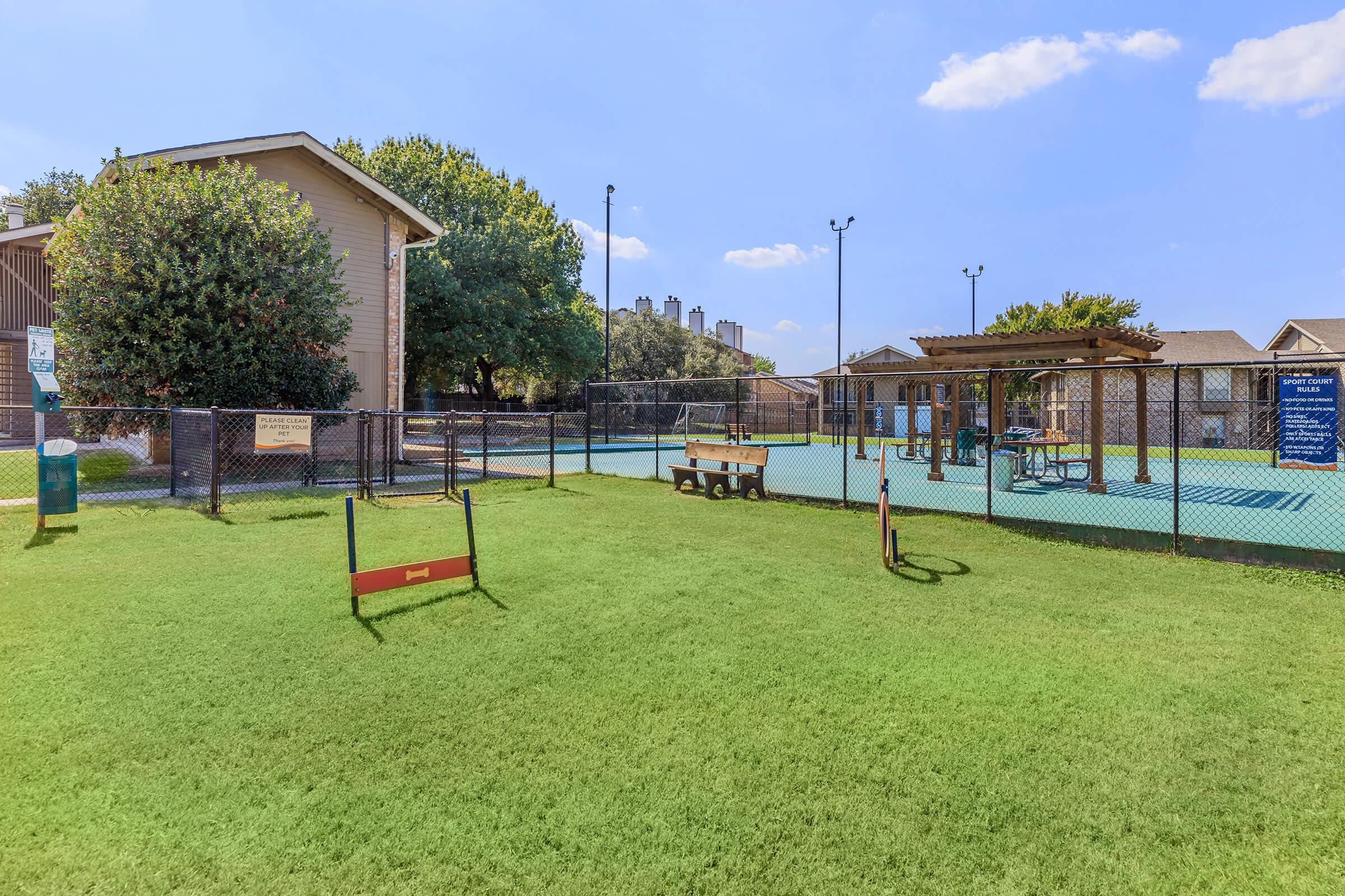 A spacious outdoor area featuring a grassy lawn, a fenced-in dog park with agility equipment, benches, and a shaded seating area. Nearby, there are amenities like a basketball court and picnic tables, surrounded by trees and residential buildings under a blue sky.