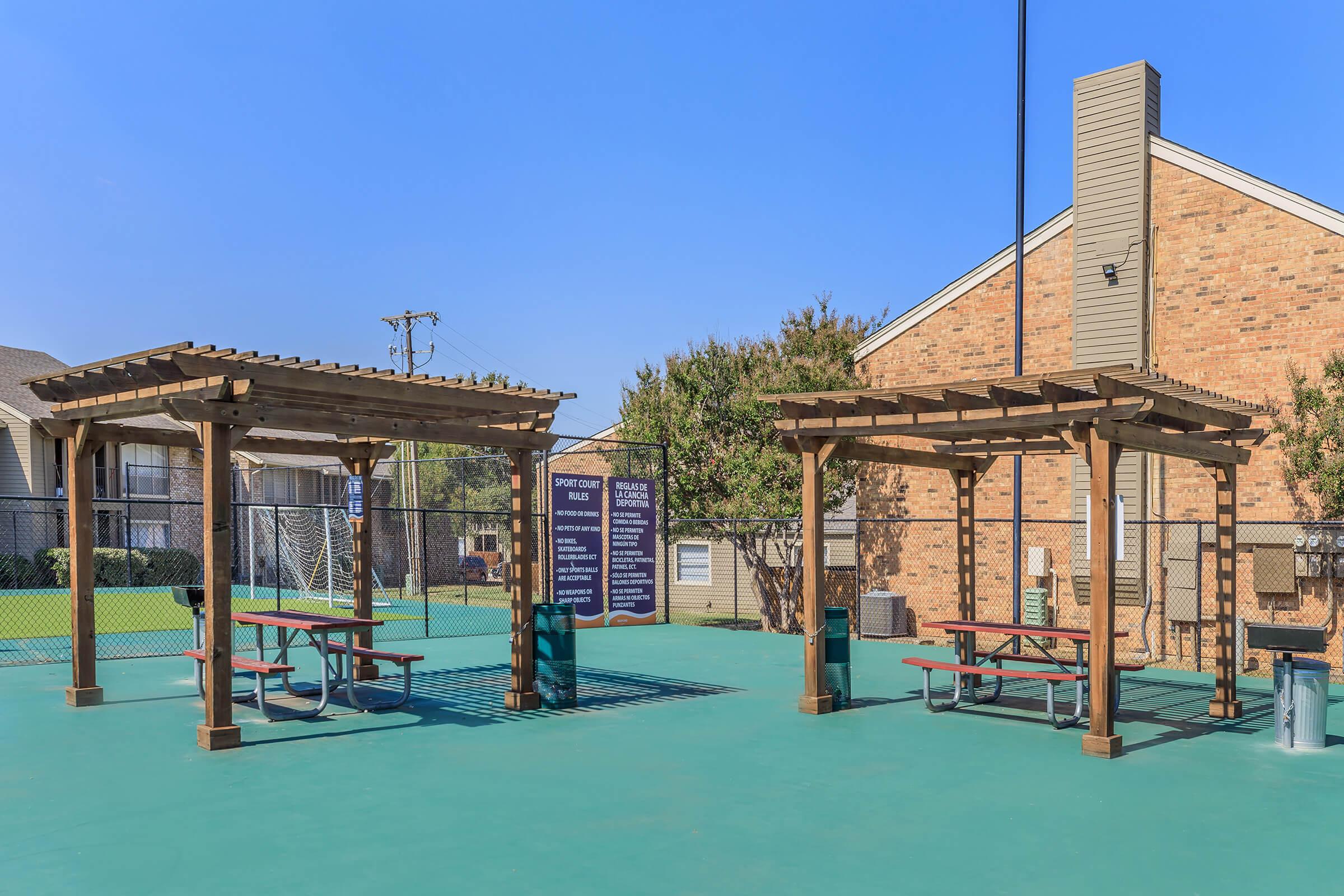Two wooden pavilions with picnic tables under them are situated on a green court. A fence encloses the area, and there is a light pole in the center. In the background, a brick building and additional recreational facilities are visible, with clear blue skies above.