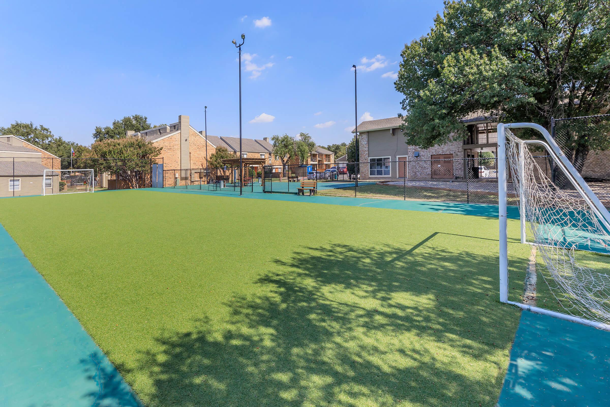 A grassy soccer field with a goalpost on one end, surrounded by a fence. The field is lined with trees and has a clear blue sky overhead. In the background, there are residential buildings visible. The scene is well-lit and inviting for outdoor sports activities.