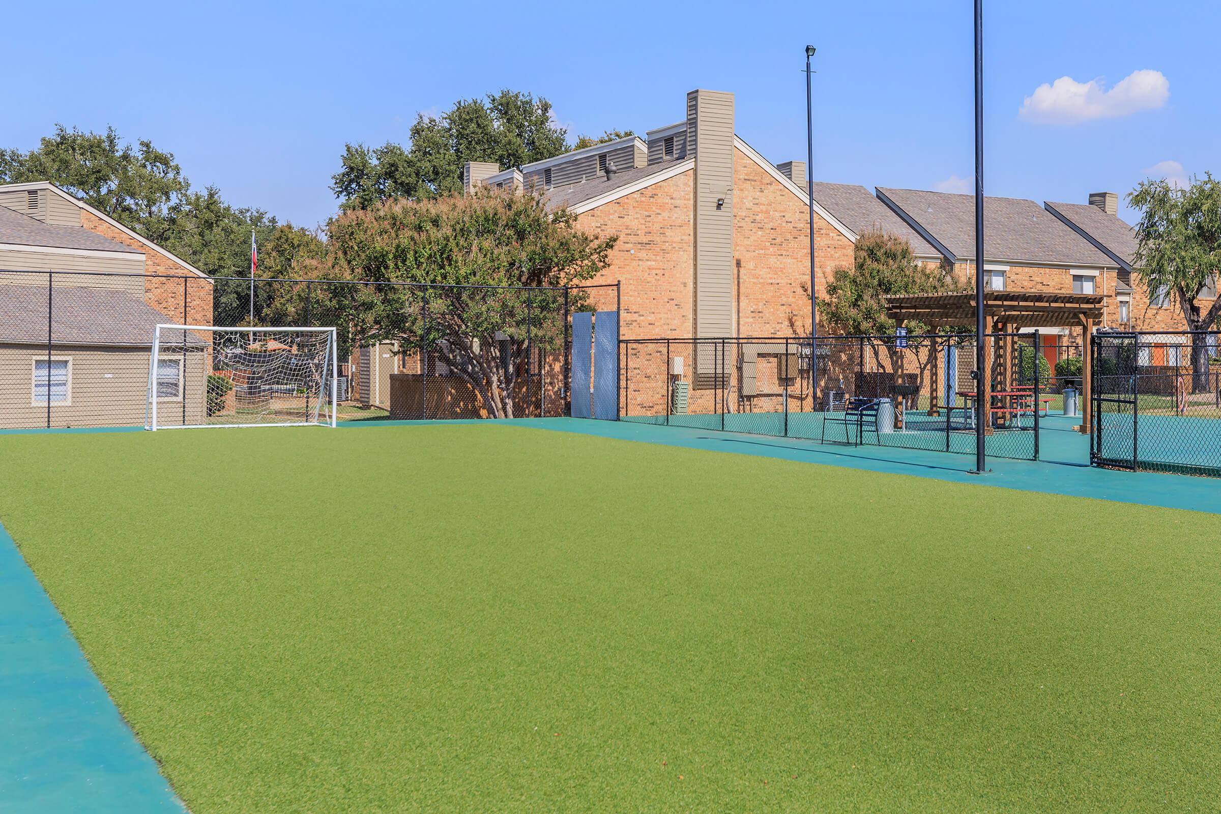 Soccer field with artificial grass in a residential area, featuring a goalpost, surrounded by brick buildings and trees. A playground and other structures are visible in the background under a clear blue sky.