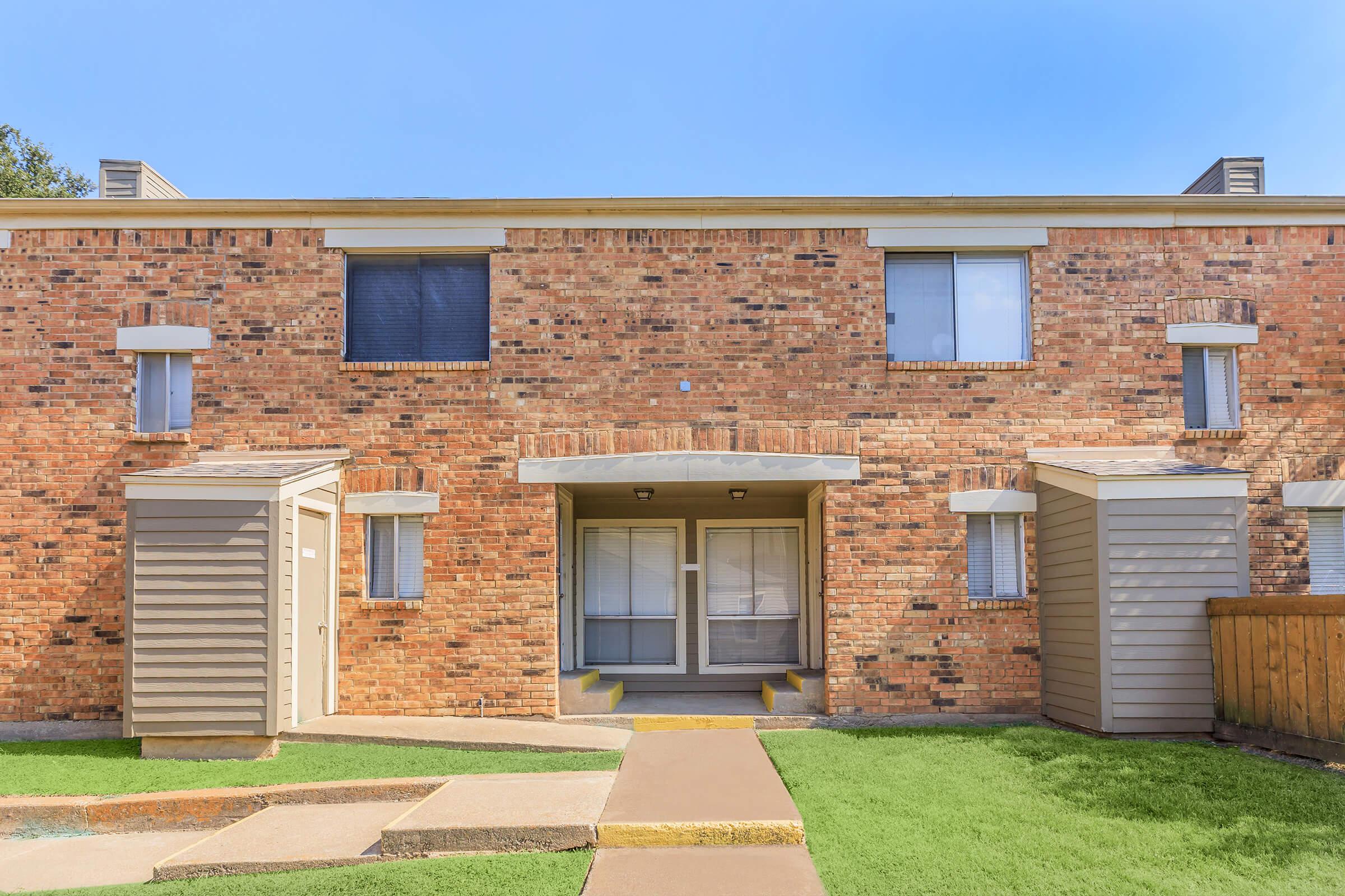 Two-story brick apartment building with two entrance doors. The facade features multiple windows with light-colored shutters. In the foreground, there's a small landscaped area with green grass and a concrete walkway leading to the entrances. Clear blue sky above.