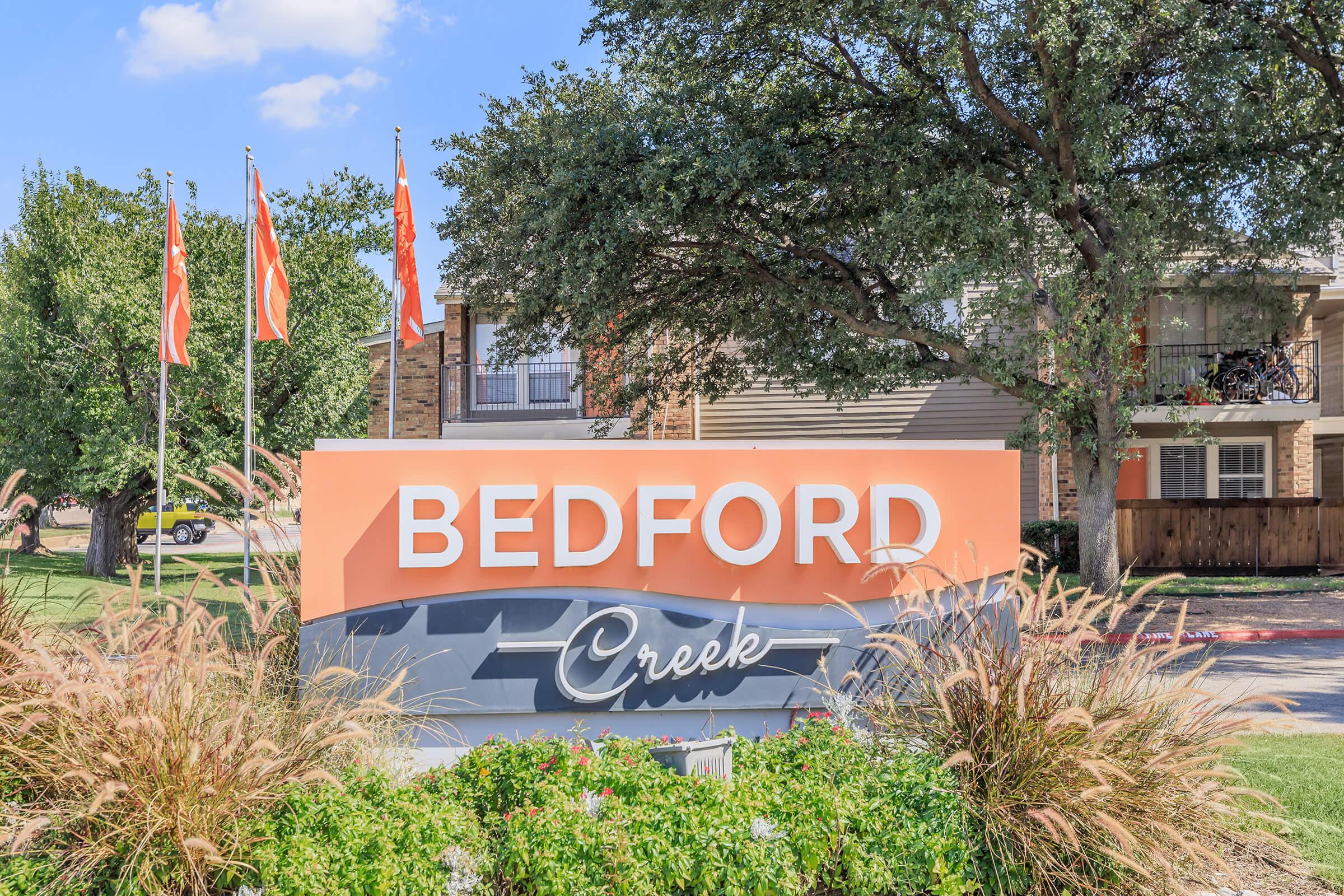 A large sign displaying "BEDFORD Creek" at an apartment complex, surrounded by landscaping and grass. Flags in the background are fluttering, and there are trees providing shade nearby. The sky is clear with a few clouds.