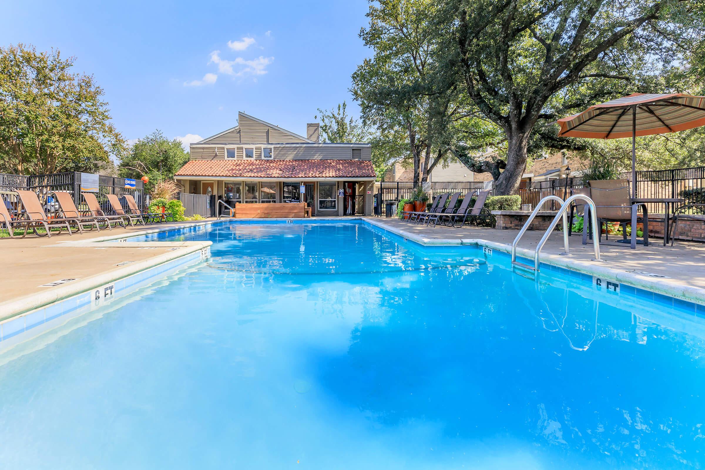 Outdoor swimming pool with clear blue water, surrounded by lounge chairs and shaded areas. A building with a tiled roof is visible in the background, along with trees providing shade. The scene is bright and inviting, ideal for relaxation and leisure activities.