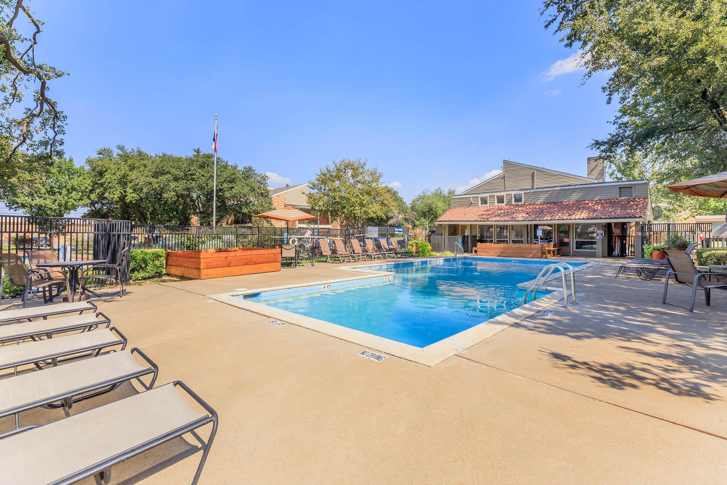 A sunny outdoor swimming pool surrounded by lounge chairs and landscaped greenery. In the background, there are trees, a building with a covered seating area, and a flagpole. The clear blue sky adds to the inviting atmosphere of this recreational space.