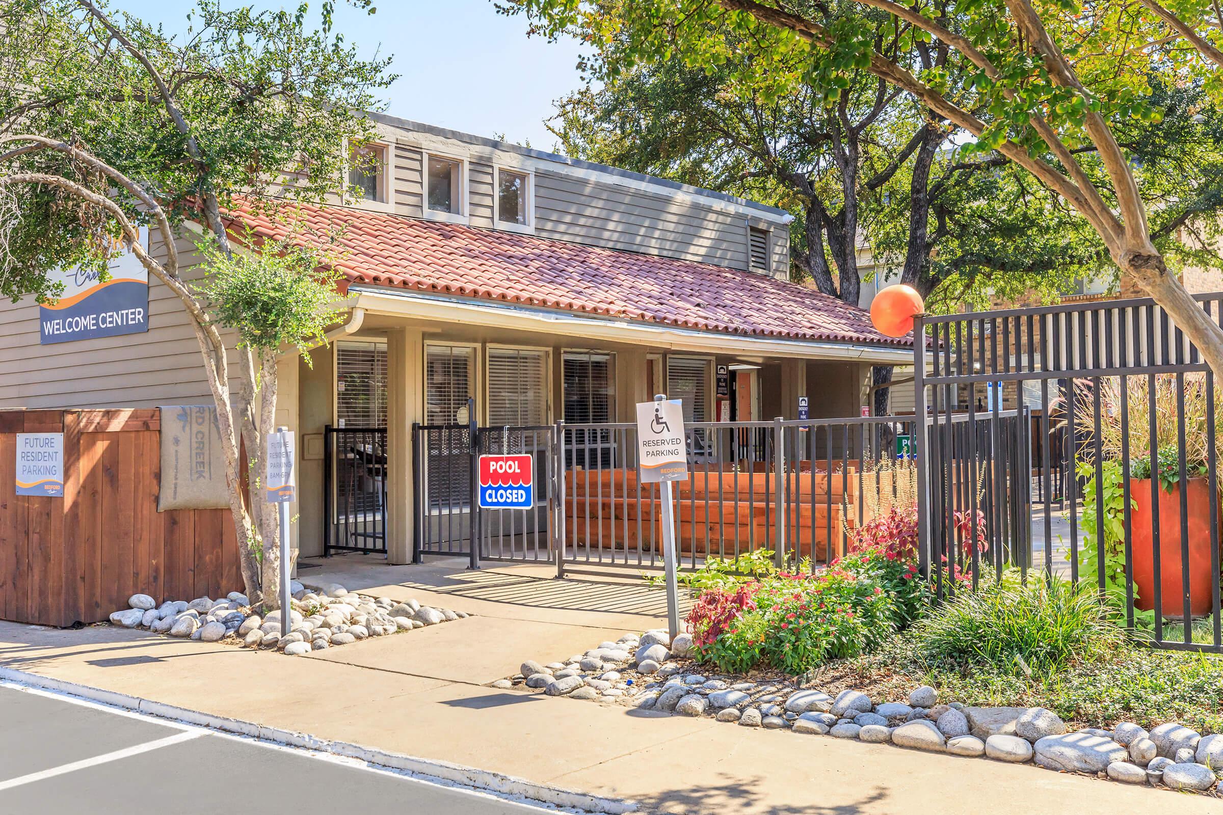 A well-lit welcome center with a Spanish tiled roof, surrounded by greenery. There is a gated entrance with signs indicating "Welcome Center" and "Pool Closed." A pathway leads to the building, which features large windows and a porch area with seating. The setting is peaceful and inviting.