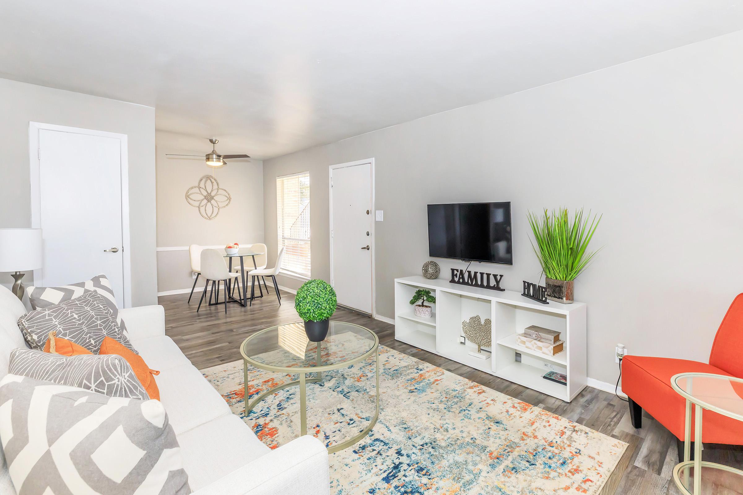 A bright and modern living room featuring a white sofa with decorative pillows, a round glass coffee table, and a colorful area rug. A TV is on a white entertainment unit, and there's a potted plant beside it. A red armchair adds a pop of color. The space includes a dining area with a small table and chairs in the background.